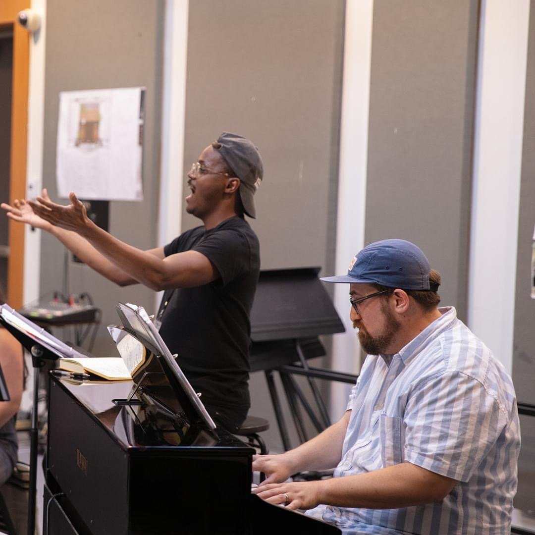 Ty leads a vocal coaching session at the piano, arms raised mid-phrase